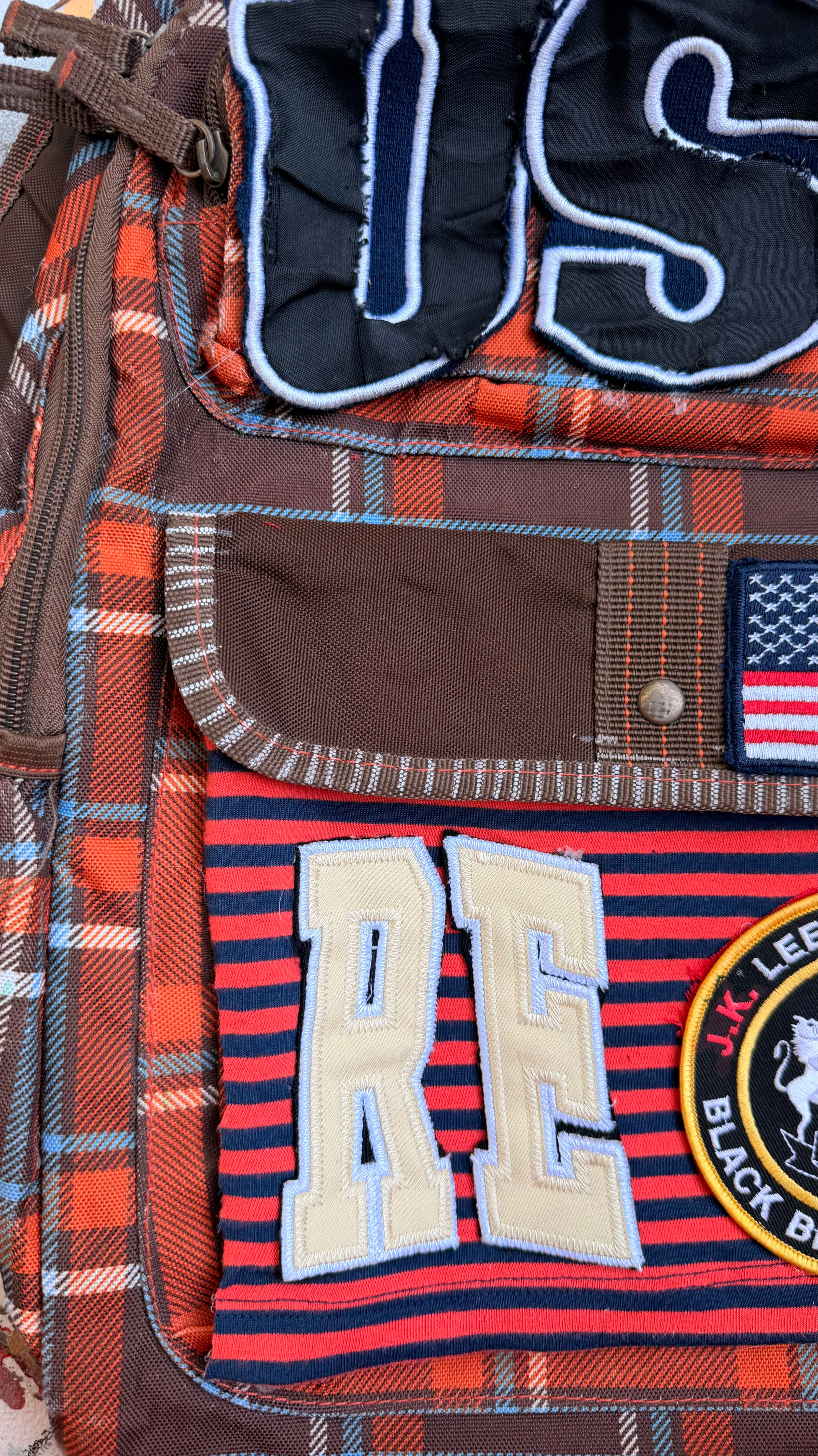 Close-up of a patchwork backpack featuring a red and blue plaid fabric, a brown flap pocket with a silver snap, a red and navy striped pocket with cream varsity-style 'RE' lettering, a small American flag patch, and a circular 'Black Belt' patch.